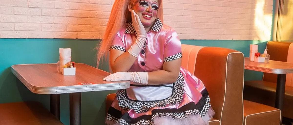 Drag queen resting a gloved hand on her cheek at a retro diner brunch table with a mimosa