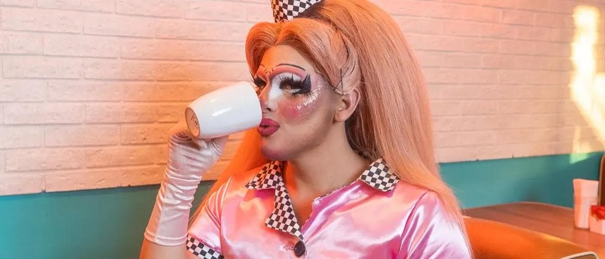 Playful drag performer sipping from a mug in a pink diner uniform at brunch