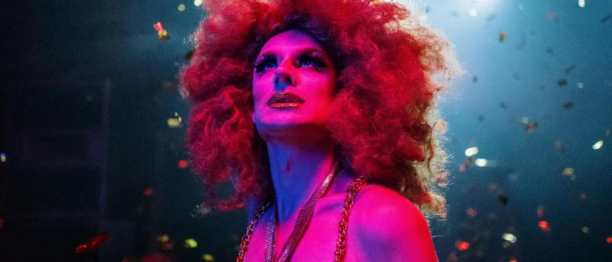 Drag queen with a red afro wig lit by pink stage light amid falling confetti at a live show
