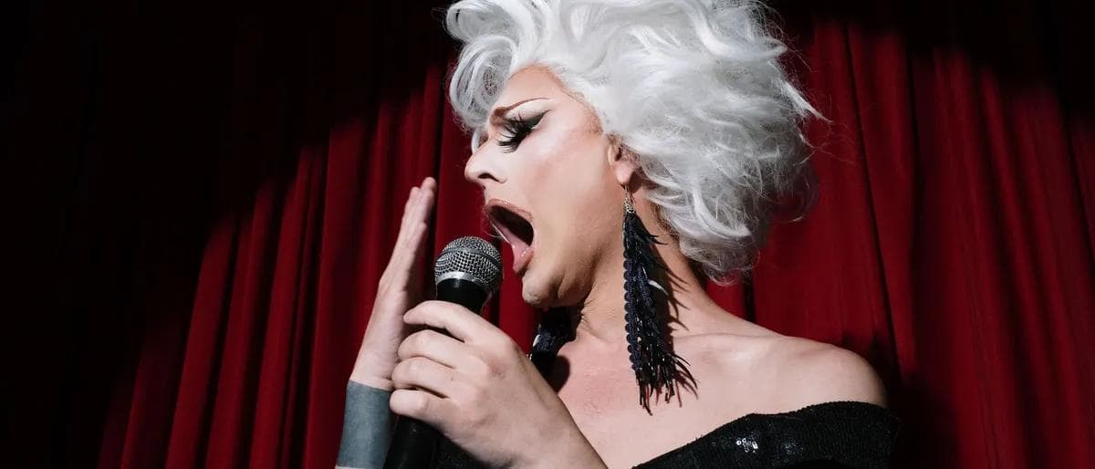 Drag queen in a silver wig singing into a microphone against a red stage curtain