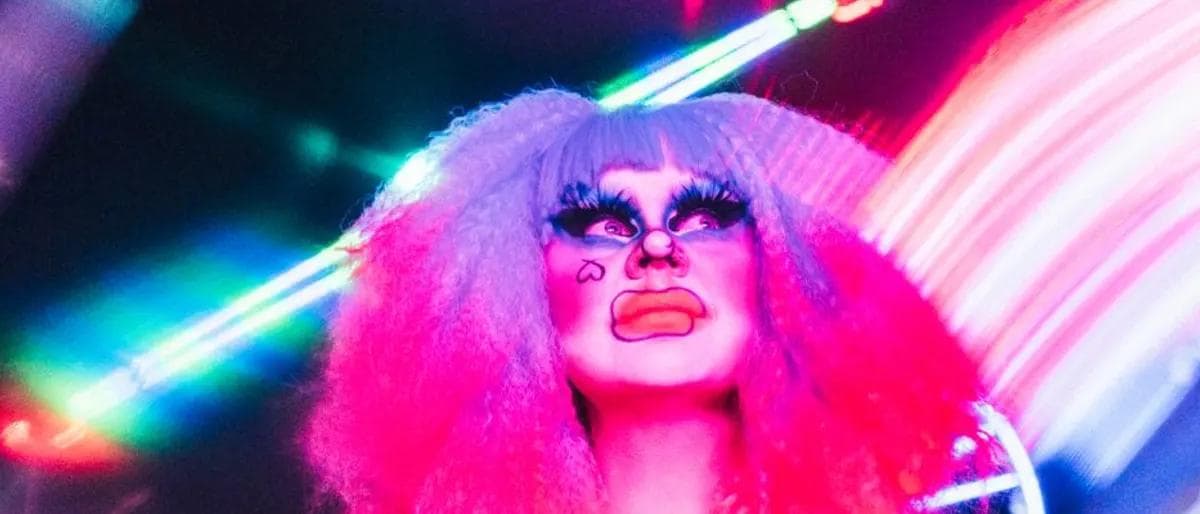 Drag performer in a pink wig under colorful laser club lights during a night show